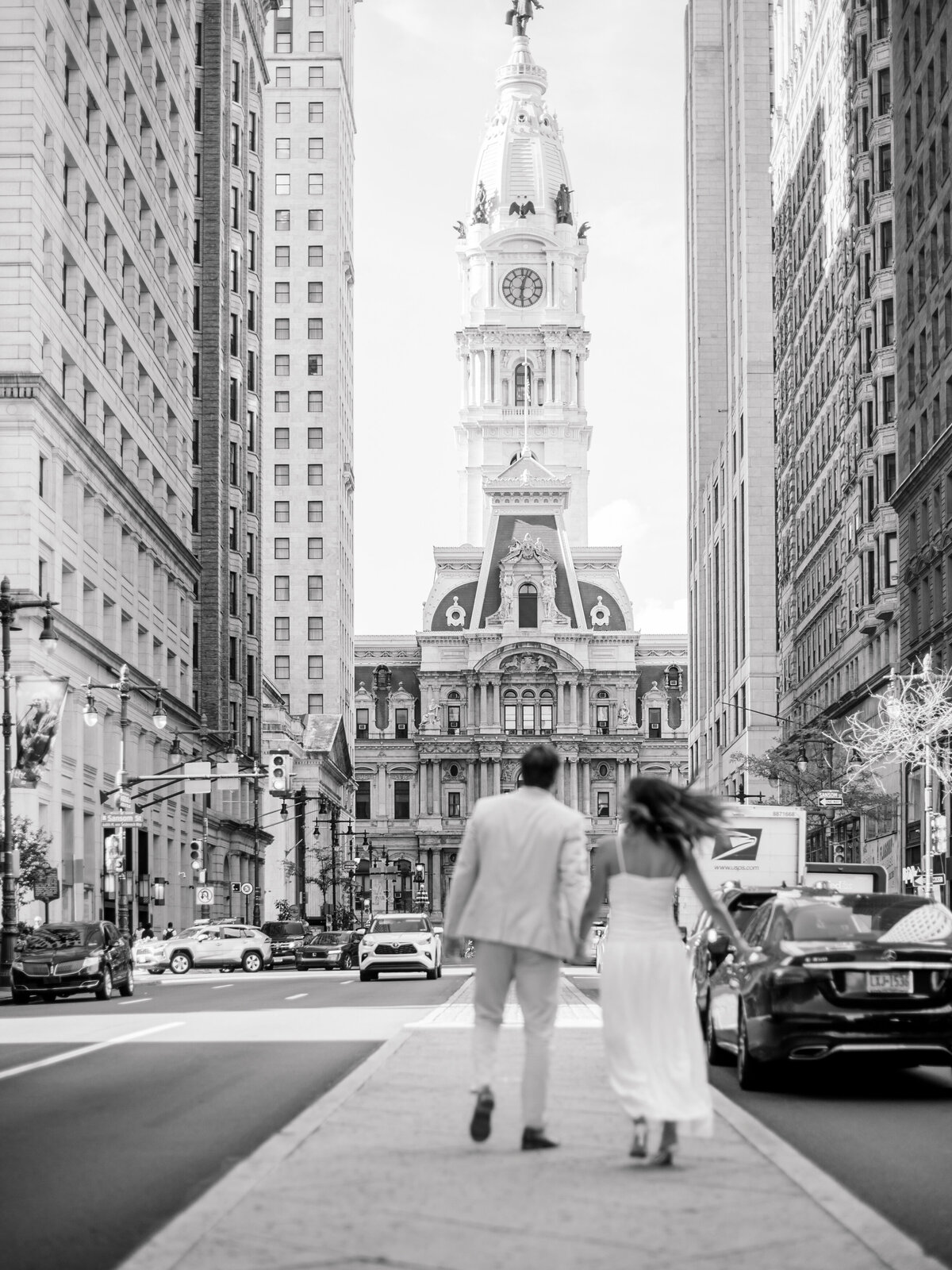 A black-and-white image of a couple walking hand in hand toward Philadelphia’s iconic City Hall along Broad Street. The groom-to-be wears a light suit, while the bride-to-be flows beside him in a white dress, her hair catching the wind. Captured by Elizabeth Kane Photography, a Luxury Philadelphia Wedding Photographer, the moment feels cinematic and timeless.