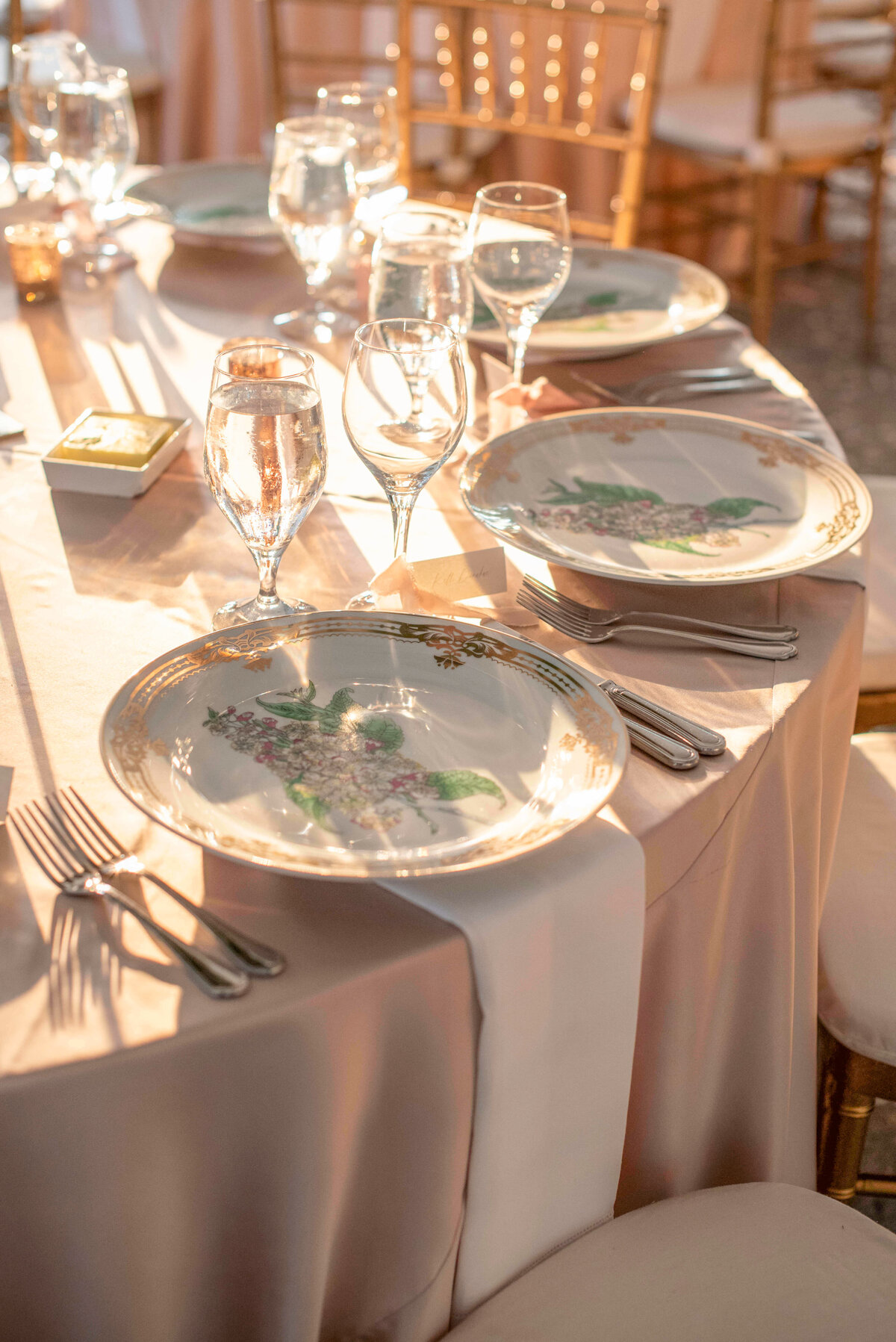 A sunlit table set for a formal dinner with elegant plates, silverware, and sparkling glassware. The round table is covered with a light pink tablecloth and surrounded by wooden chairs, creating a warm and inviting atmosphere.