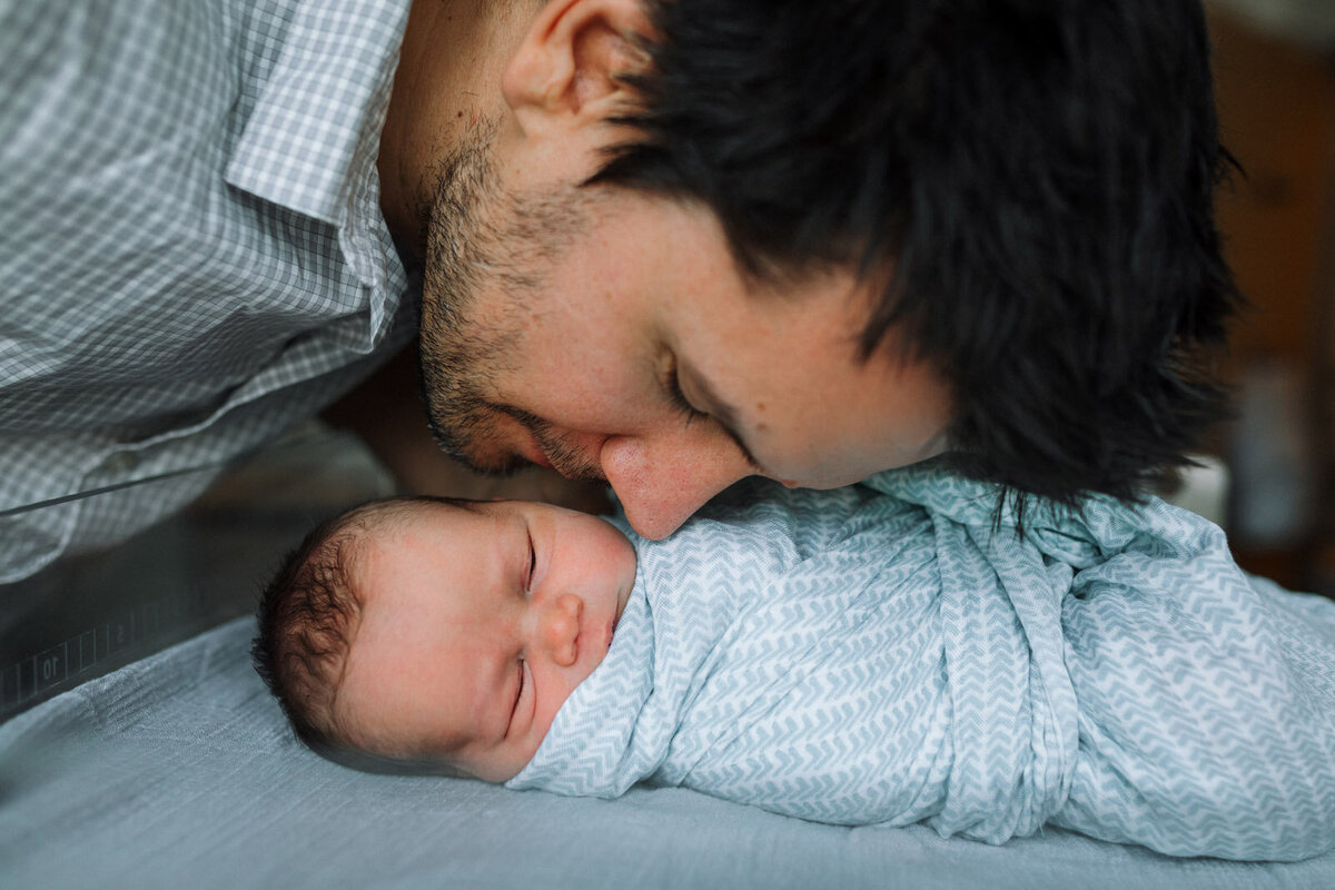 dad leaning over to swaddled newborn in blue blanket in bassinet