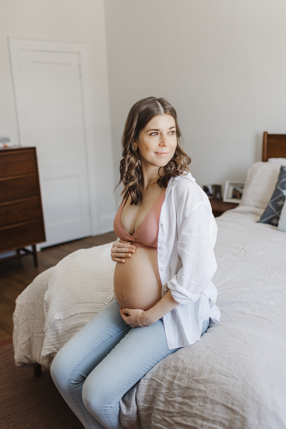 Brunette pregnant woman dressed in jeans, pink bra and white open shirt sitting on her bed looking out the window during her Toronto Maternity Photography session
