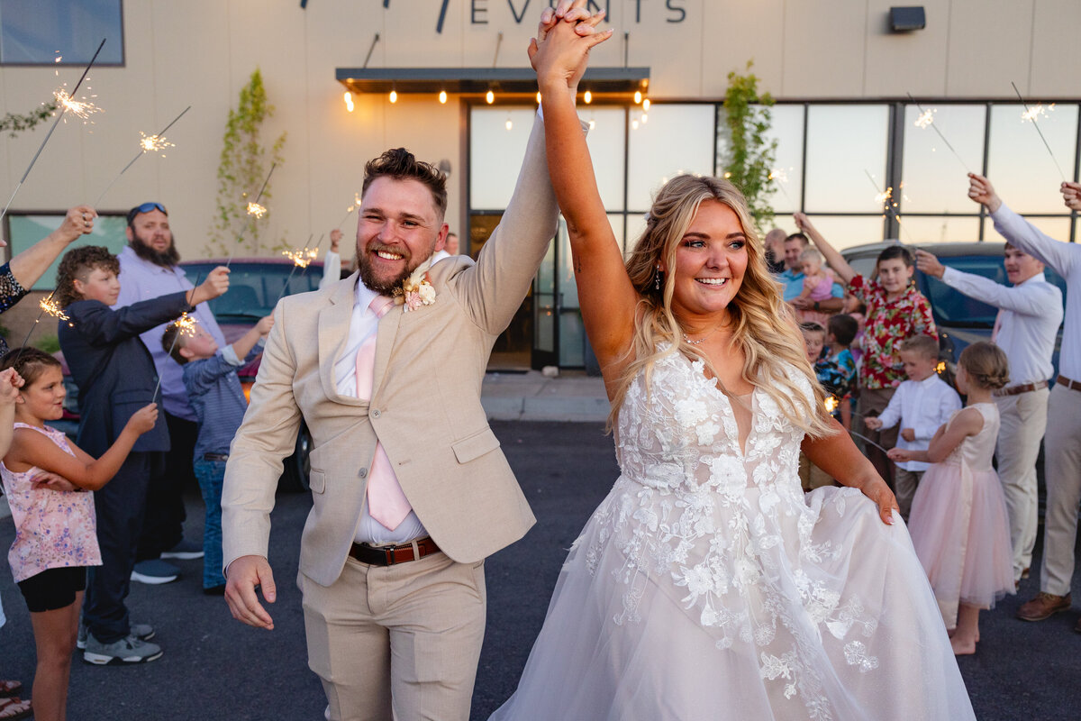 The bride and groom hold hands high in the air, smiling as they walk through a crowd of guests holding sparklers outside a modern event venue. Captured by Cassie Lee Photography