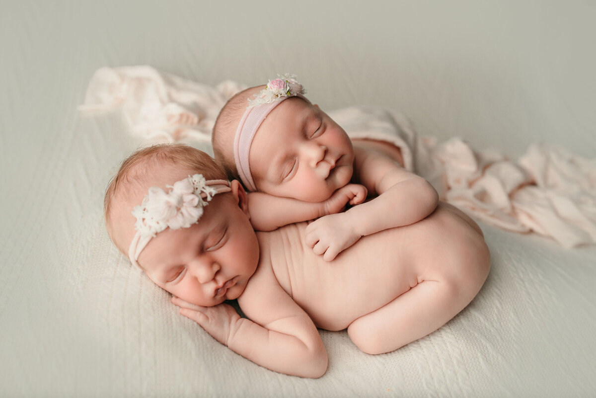 Newborn baby girl twins nude and posed laying side by side one twin is laying on the other twin's back and they have on white headbands, draped in cream fabric