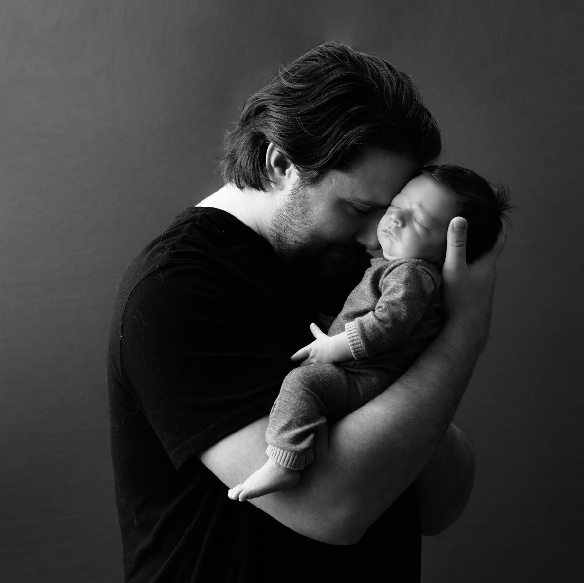 Black and white image of a new dad holding his brand new son in his arms. Dad's head is toughing baby's forehead. Image by Beth Benecki of E. Benecki Photography.