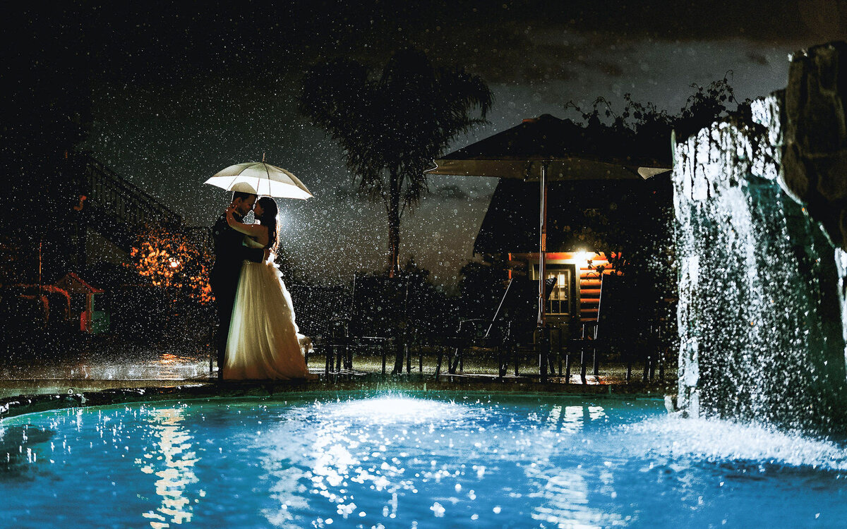 Bride and groom share a romantic moment under an umbrella in the rain, captured in a cinematic documentary style.