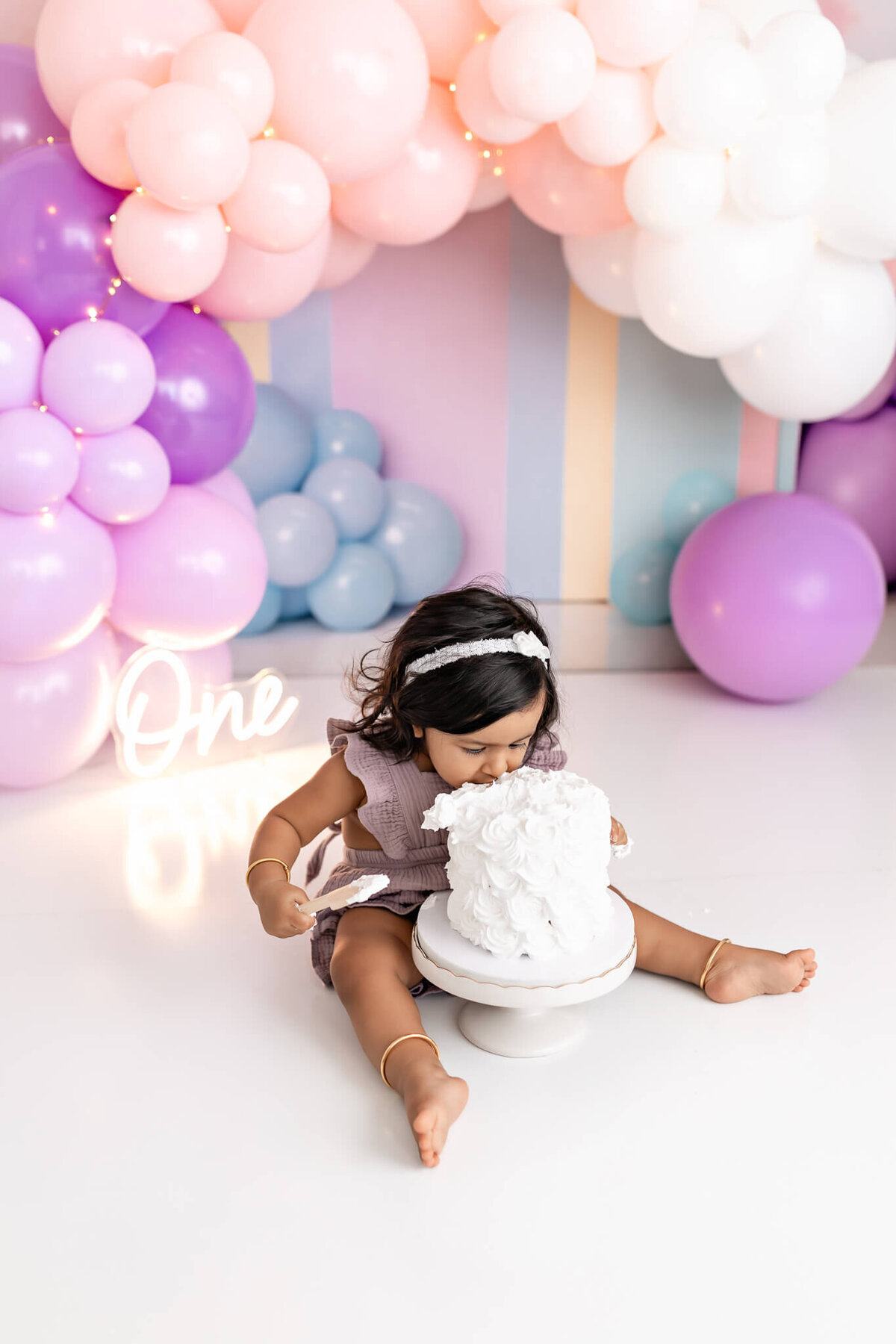 A toddler is sitting on the floor with her hands and mouth in a cake. She is surrounded by pastel balloons in pink, purple, and blue, with a neon sign that reads "One" in the background.