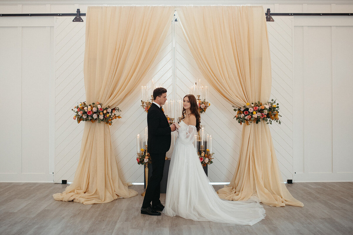 A bride and groom wearing a white wedding gown and black tuxedo stand in front of peach-colored curtains with flowers.
