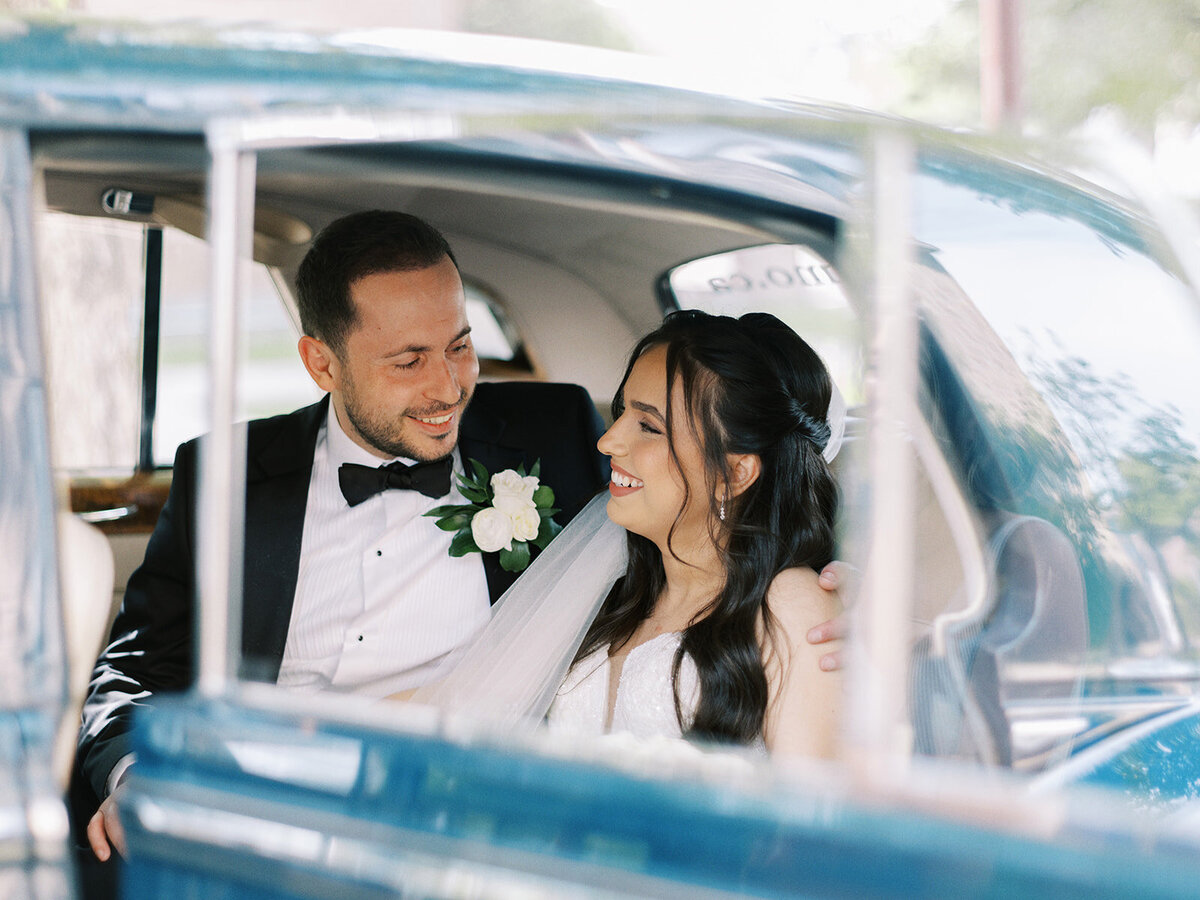 A man and woman in wedding attire are sitting together in a car, smiling at each other, after their classic Calgary wedding.