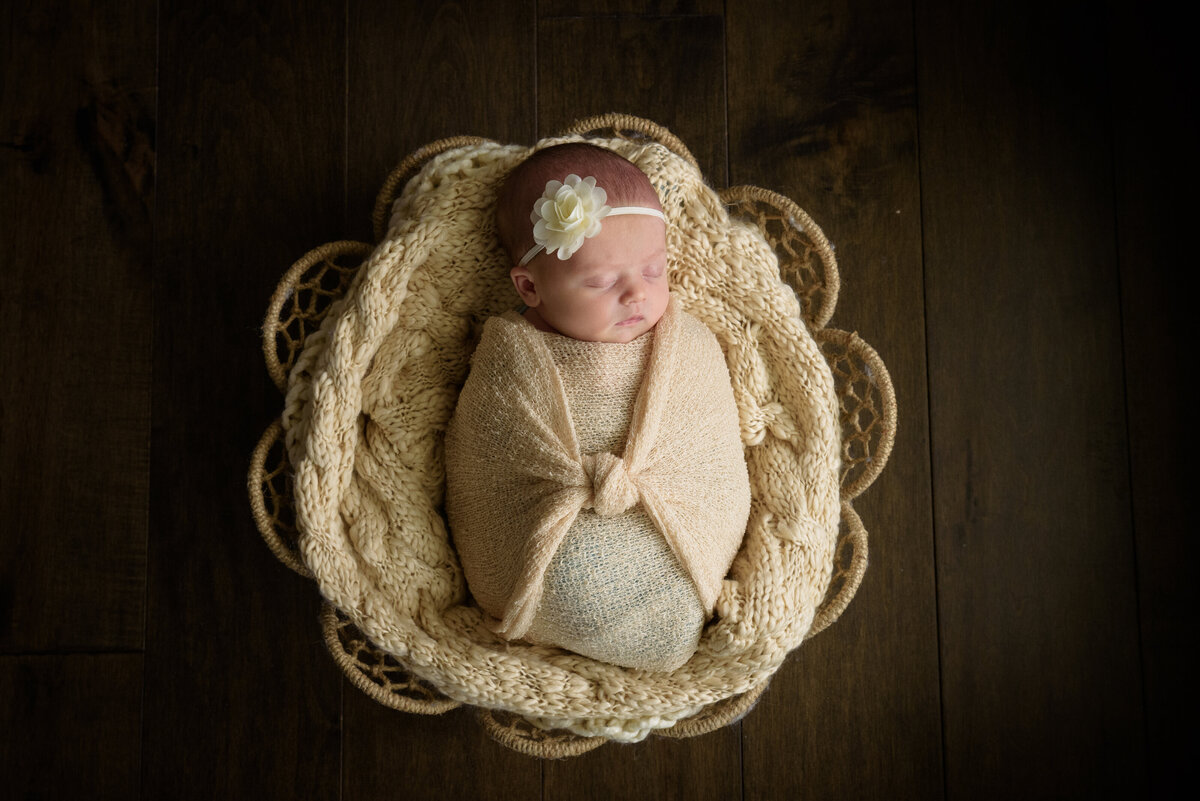 Newborn baby girl in a cream colored wrap and a cream flower posed in a basket in her home in Green Bay, Wisconsin.