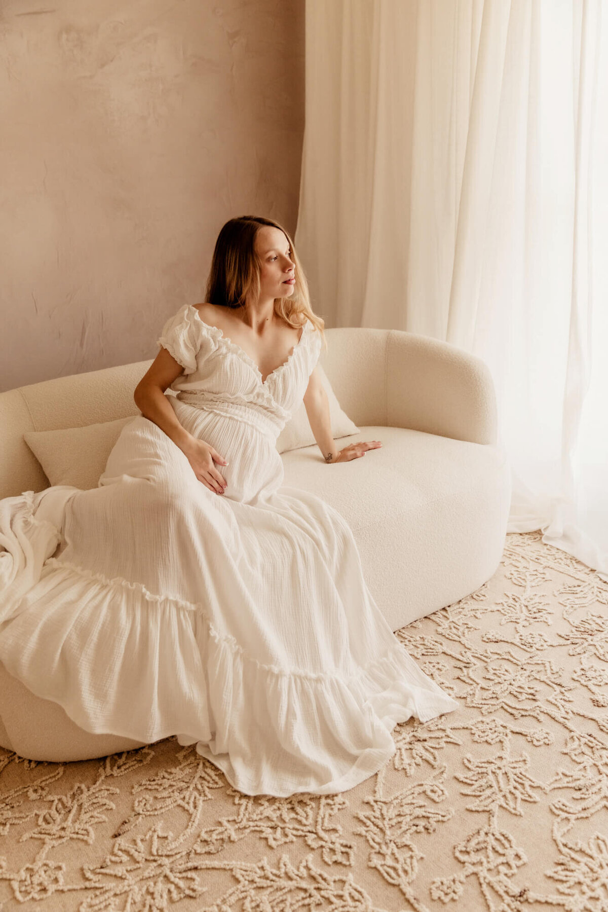 Woman wearing a white off the shoulder dress sitting on a white modern couch for a maternity portrait in Yukon, Oklahoma.