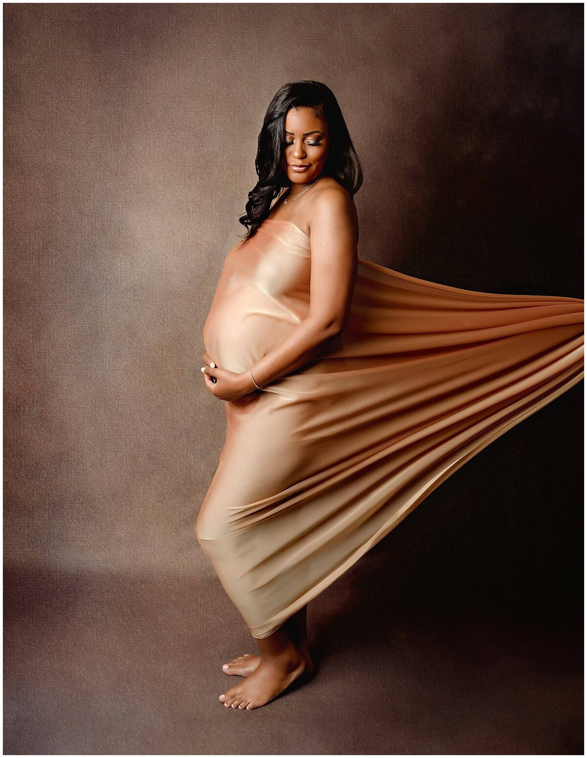 Woman holding her pregnant belly and looking down on the side while someone holds and pulls the fabric from the back creating a nice way to showcase her belly bump.