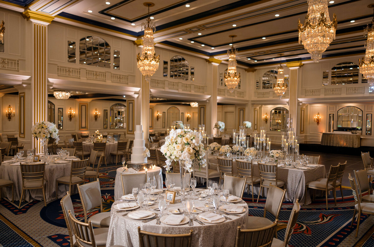 Opulent ballroom set for an event, featuring round tables with elegant white flower centerpieces and tall candles. Crystal chandeliers hang from the ceiling, illuminating the room. Tables are adorned with silverware and glassware.