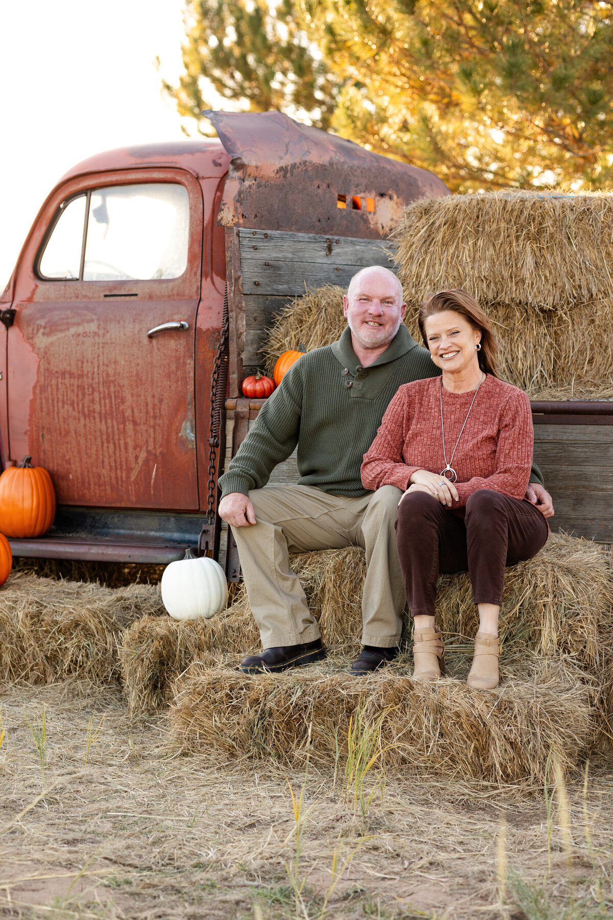 Husband and wife sit on bale of hay in front of vintage red farm truck and smile at the camera.
