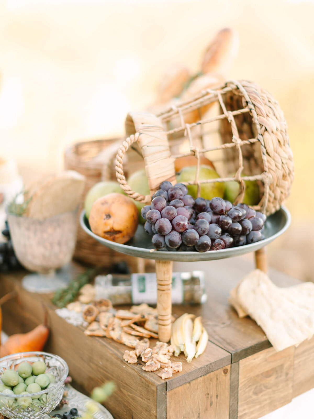 A rustic table setting with a wicker basket, green apples, and a cluster of purple grapes on a wooden stand.