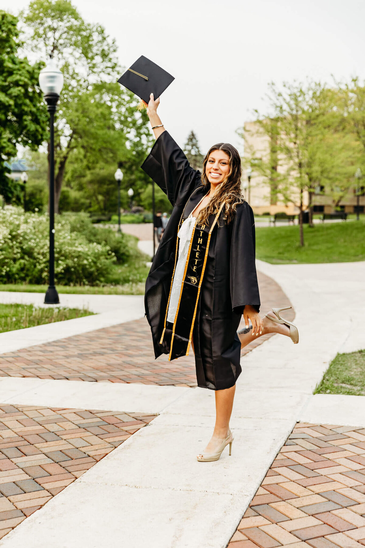 Young woman enjoying her UW Oshkosh graduation photo session as she wears her cords and gown while holding her cap in the air