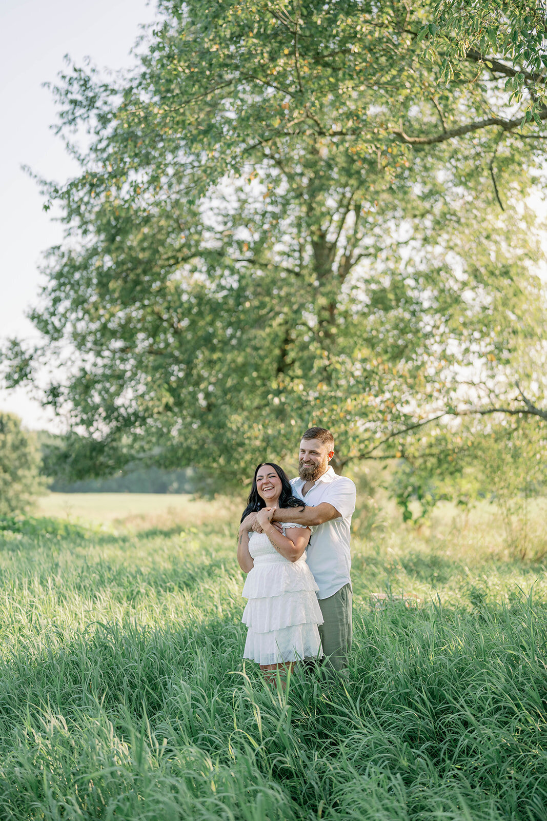 Kali and Joe hugging beneath a large tree in an open field on private property during their Detroit engagement photos.