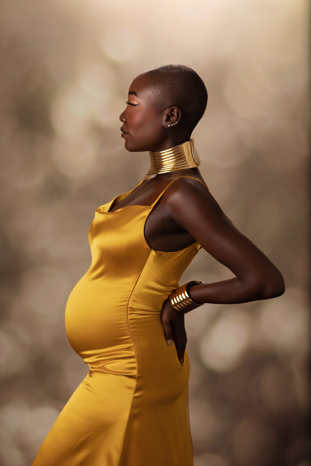 A powerful side-profile maternity photograph of an expecting mother adorned in a gold satin dress and statement gold jewelry, standing gracefully against a soft, glowing background. Captured by a skilled DFW maternity photographer.