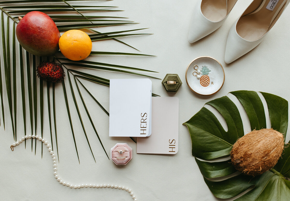 A flat lay wedding arrangement with shoes, tropical fruits, and 'His and Hers' items on a white surface with palm leaves