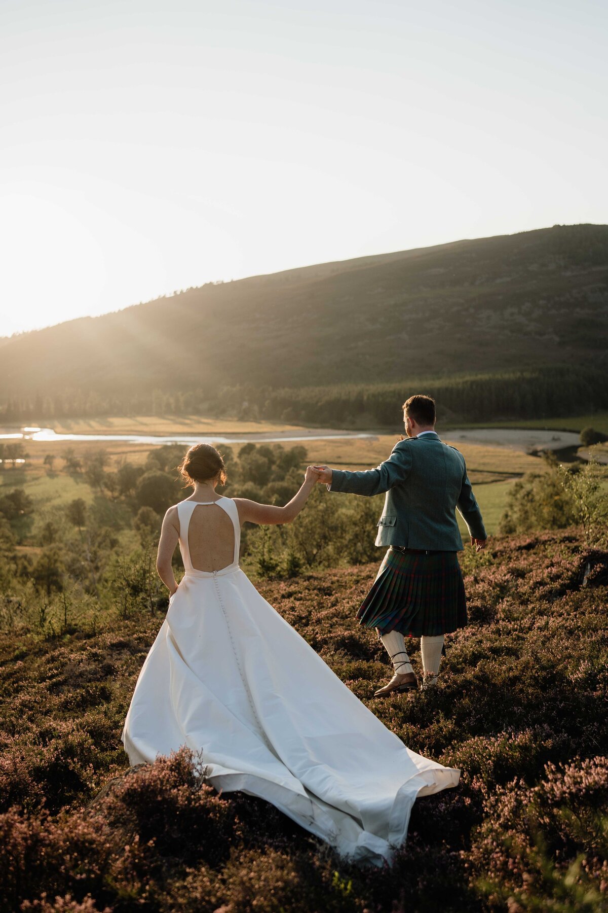 A wide photograph of a wedding couple walking hand in hand down a hill. It' golden hour and the Mar Lodge valery can be seen all around.