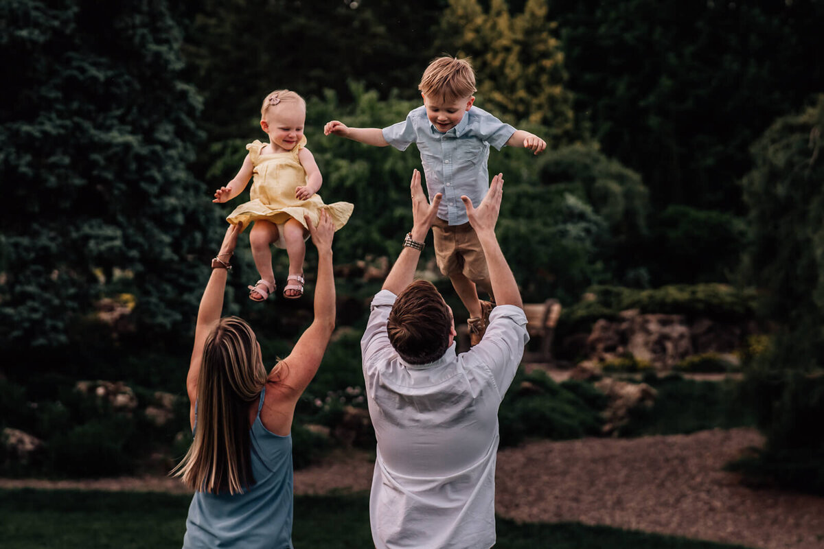 Two parents stand with their backs to the camera, gently tossing their two children up into the air at Lake Harriet Peace Gardens.