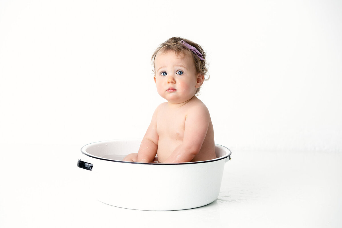 A baby girl is sitting in a white tub after her cake smash photo session.