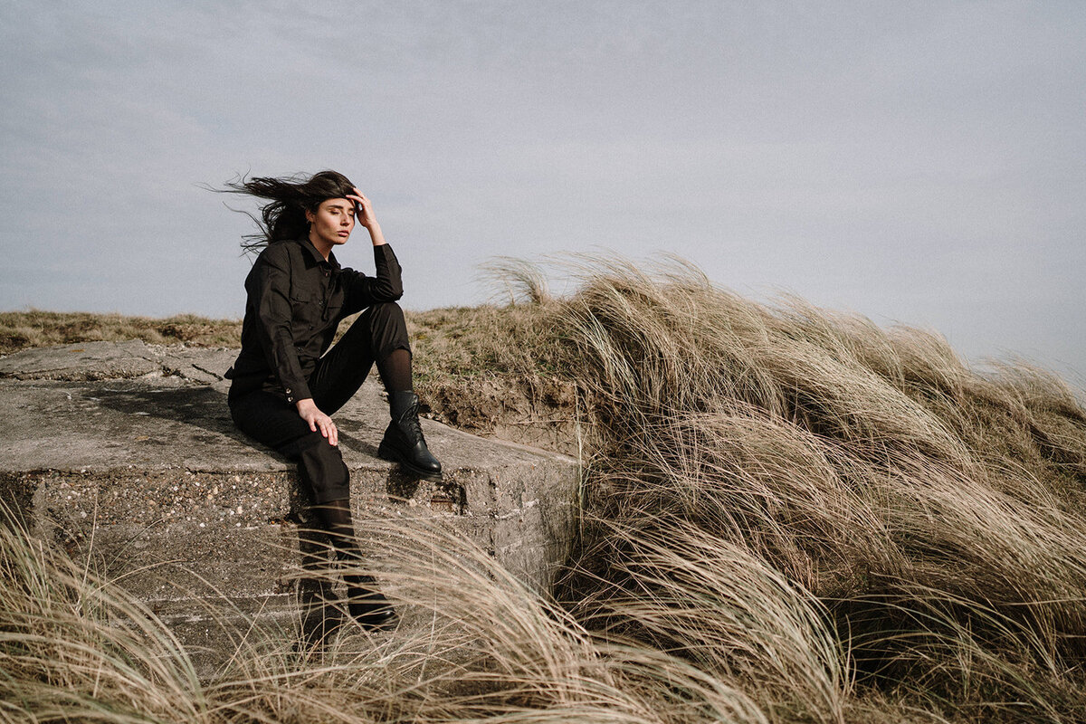 vrouw zittend in de duinen met harde wind, donkere kleding, strand portret