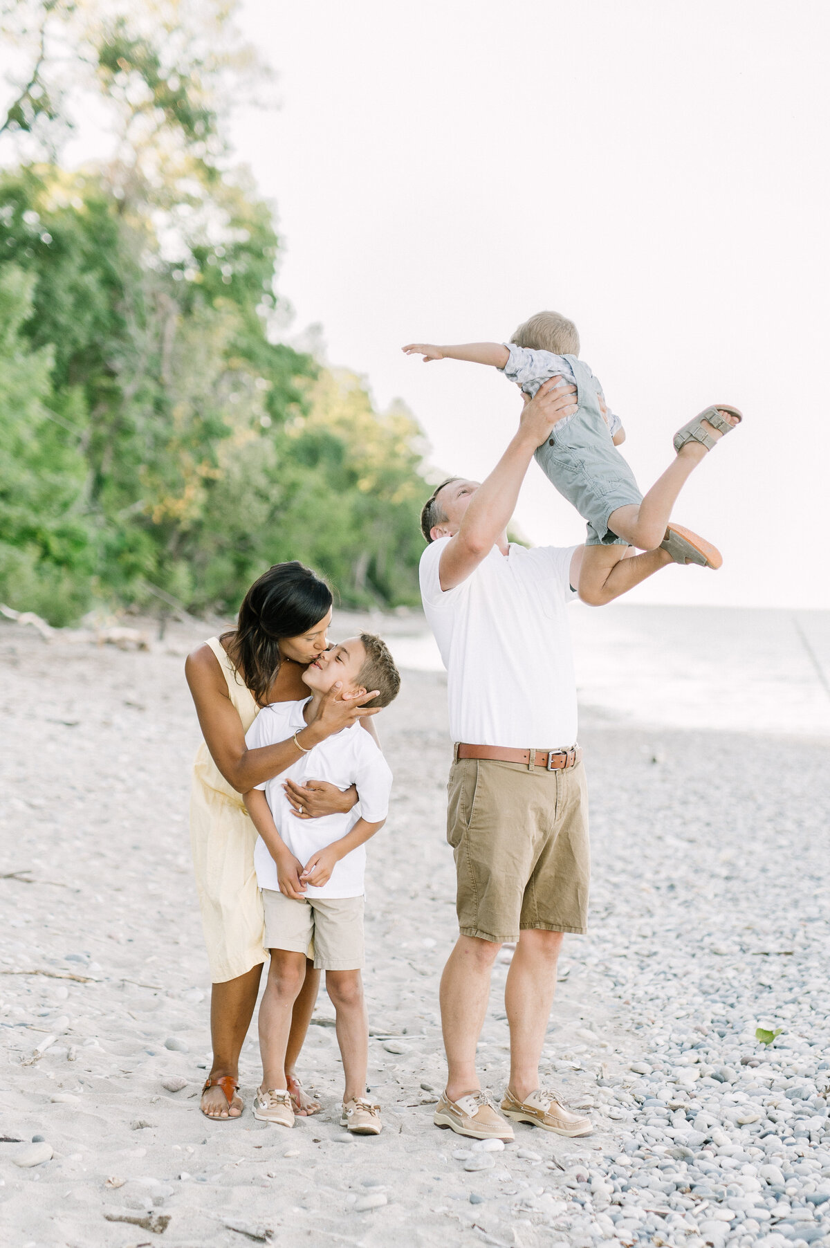 A mom is kissing her son on the beach while the dad is tossing the other child in the air