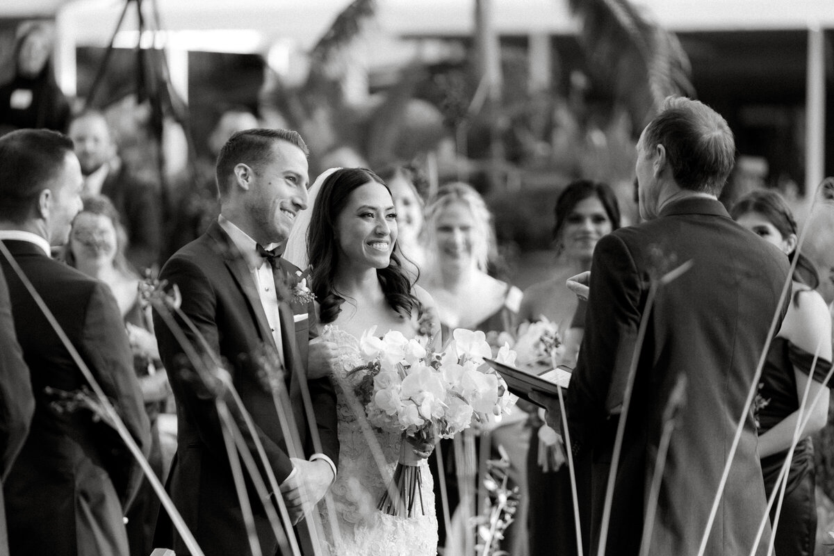 A candid black and white shot of a bride and groom smiling during their wedding ceremony, with guests and arches in the background.