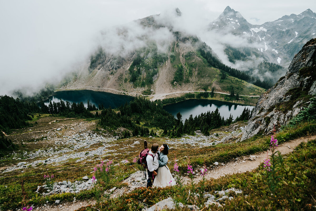 a couple embraces on a hiking trail lined with blooming Alaskan fire weed. Their adventure elopement photographers shoot from the switchback above them giving a sweeping perspective of the mountains and alpine lakes that surround them. 