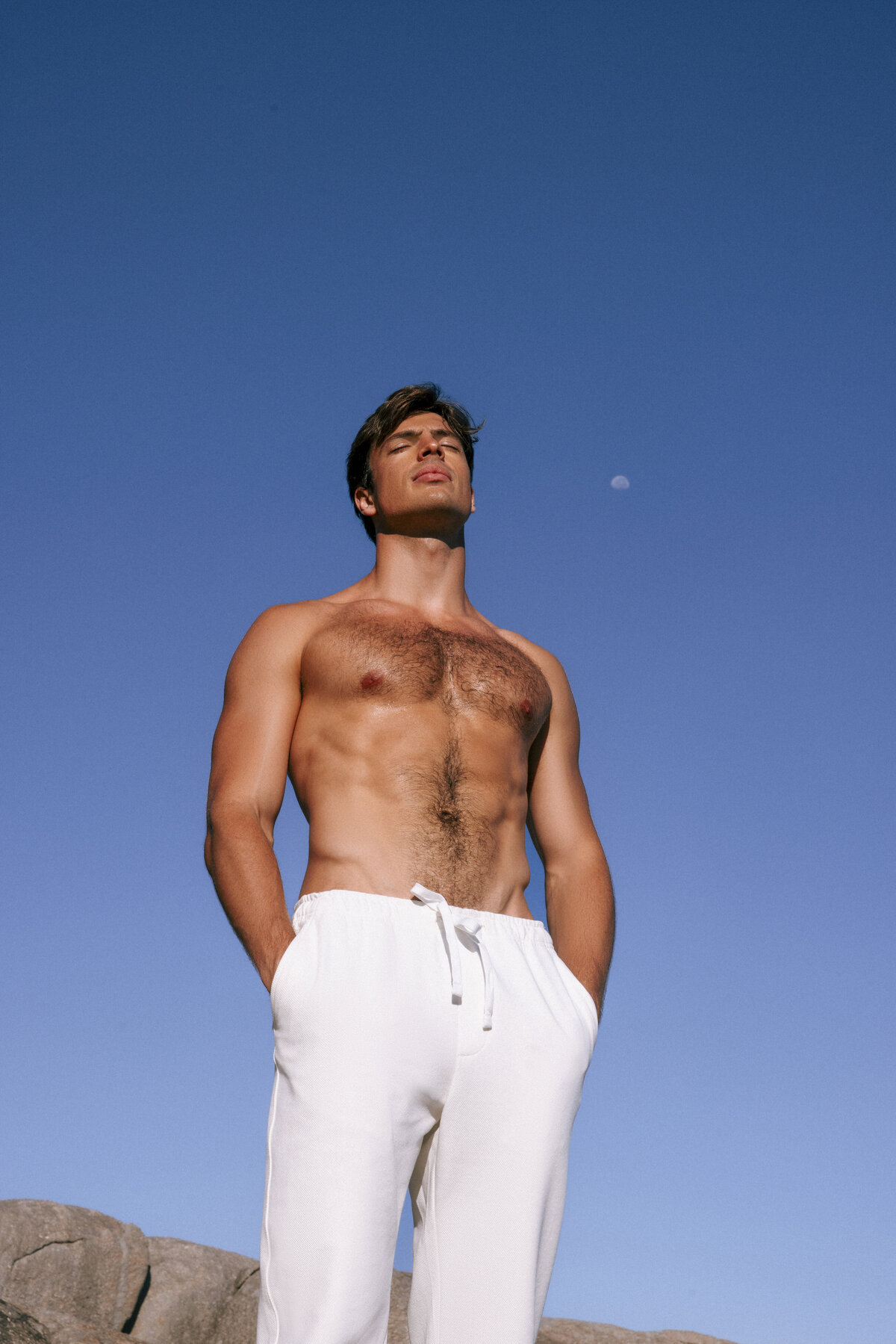 Shirtless male model standing confidently with eyes closed under the clear Cape Town sky, wearing white drawstring pants. A serene and powerful portrait with the moon subtly visible in the background.