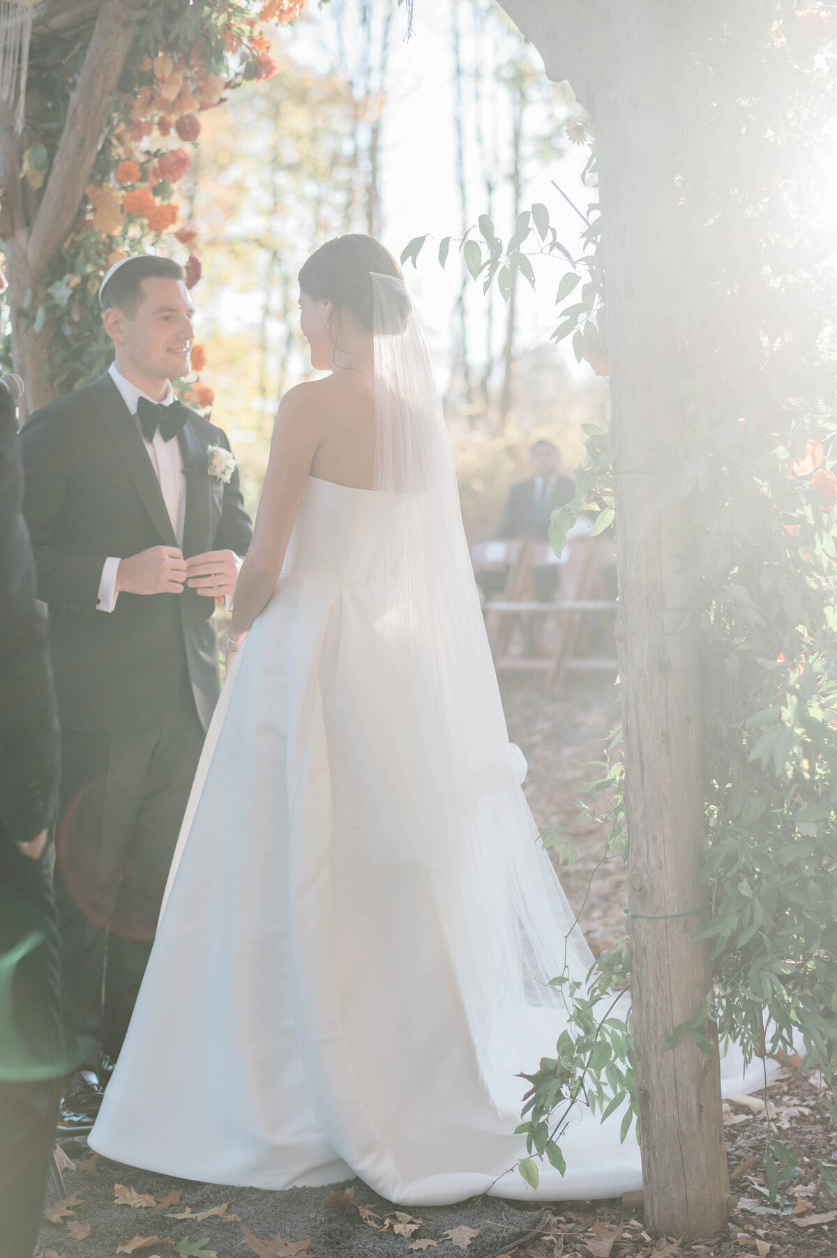 The bride and groom stand beneath the floral-covered chuppah, bathed in warm, golden light