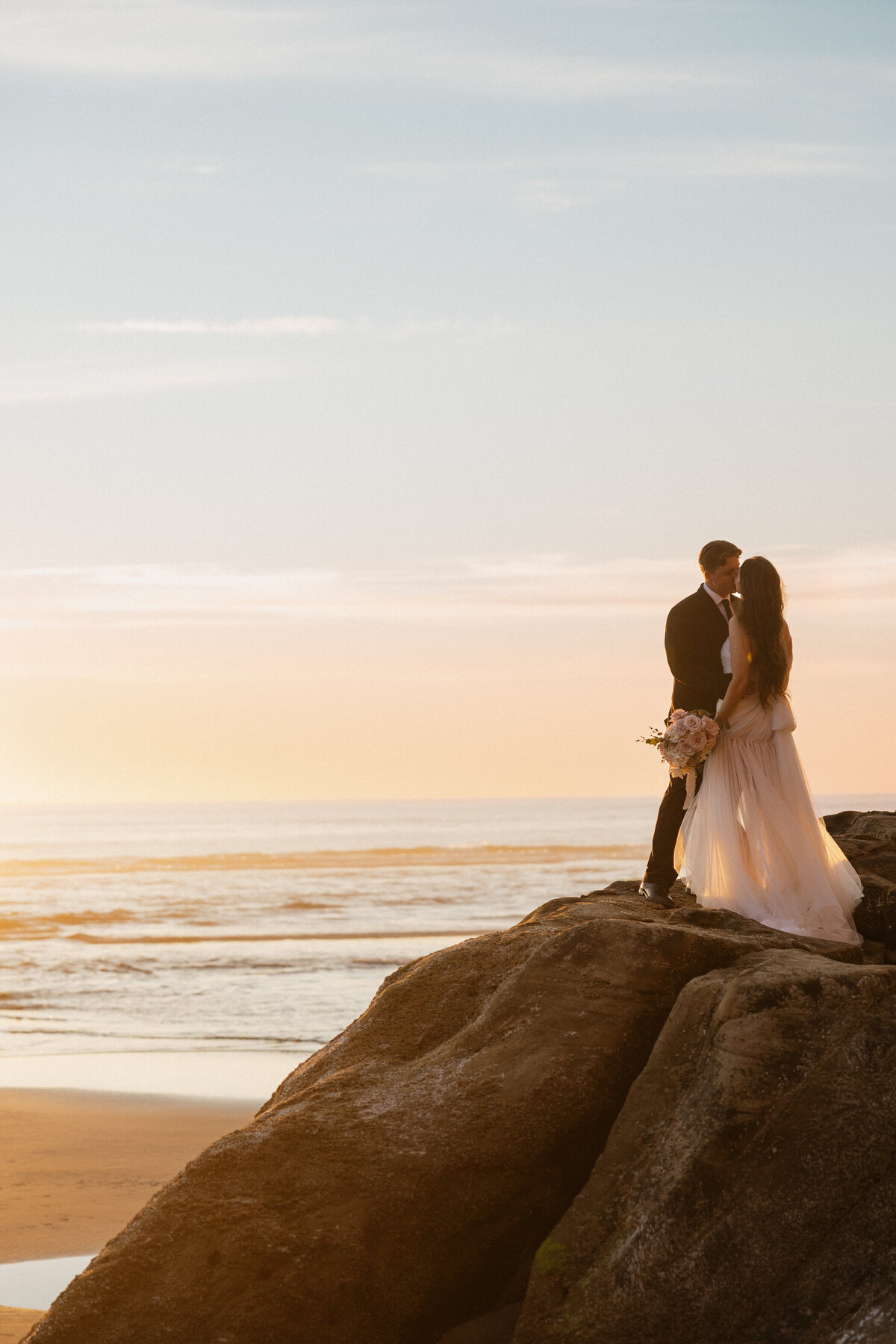 Bride and groom stand silhouetted on a rocky cliff at sunset in Oregon