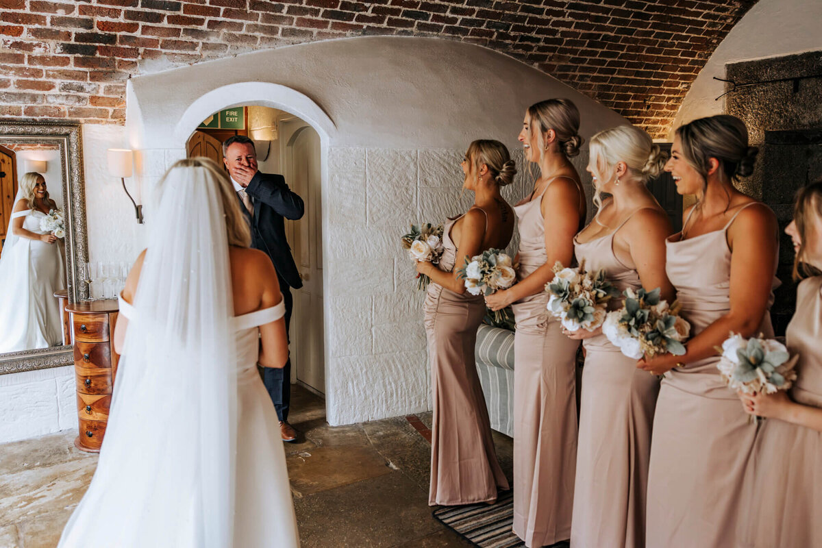 A line of bridesmaids on one side, the bride on the other and the brides father in the doorway looking at the bride with his hand covering his mouth in shock because the bride looks so beautiful