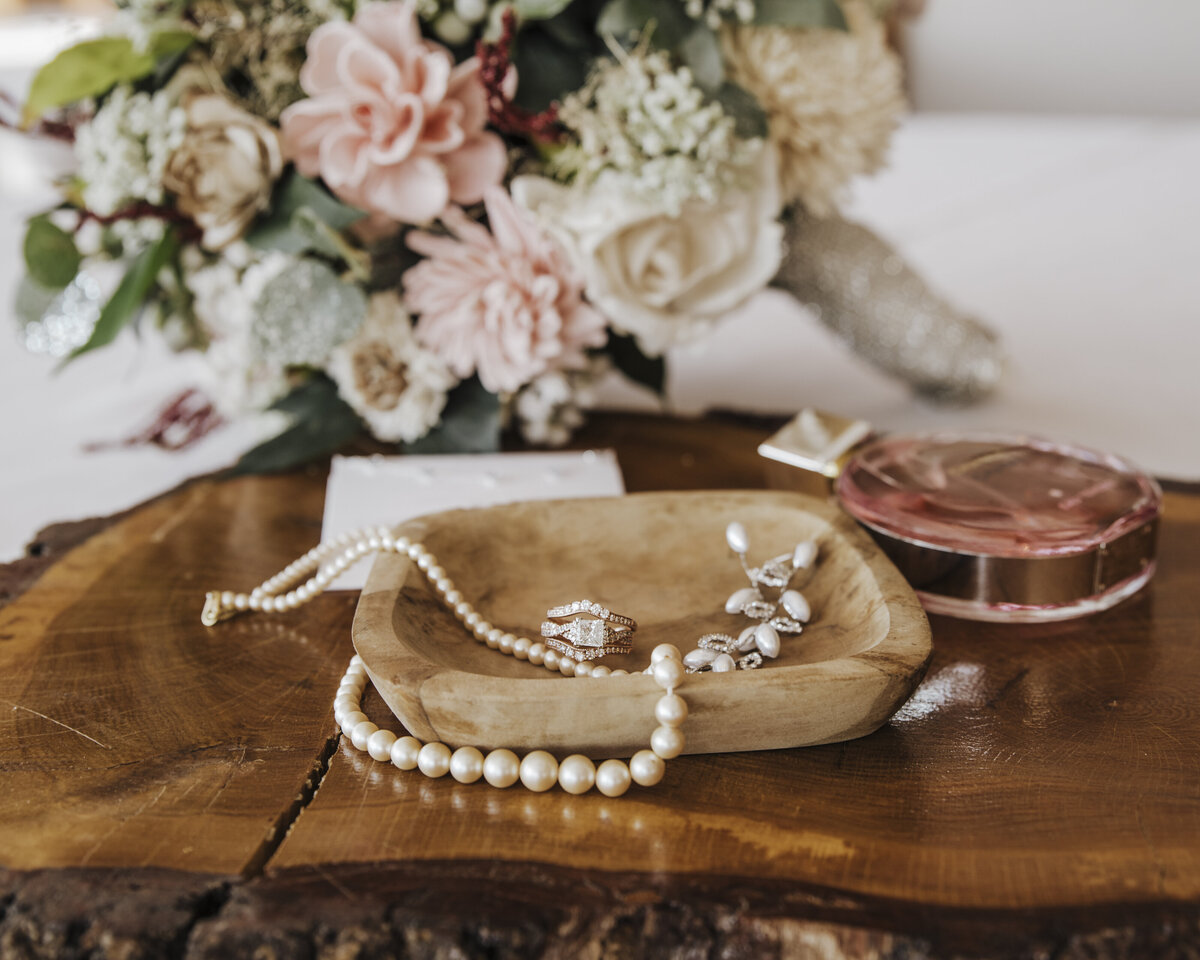 An elegant selection of bridal accessories including a string of pearls, a bracelet, and earrings meticulously placed on a wooden dish beside fresh floral arrangements and a hint of perfume, capturing the essence of a bride's preparation on her wedding day taken by jen Jarmuzek photography a Minneapolis wedding photographer