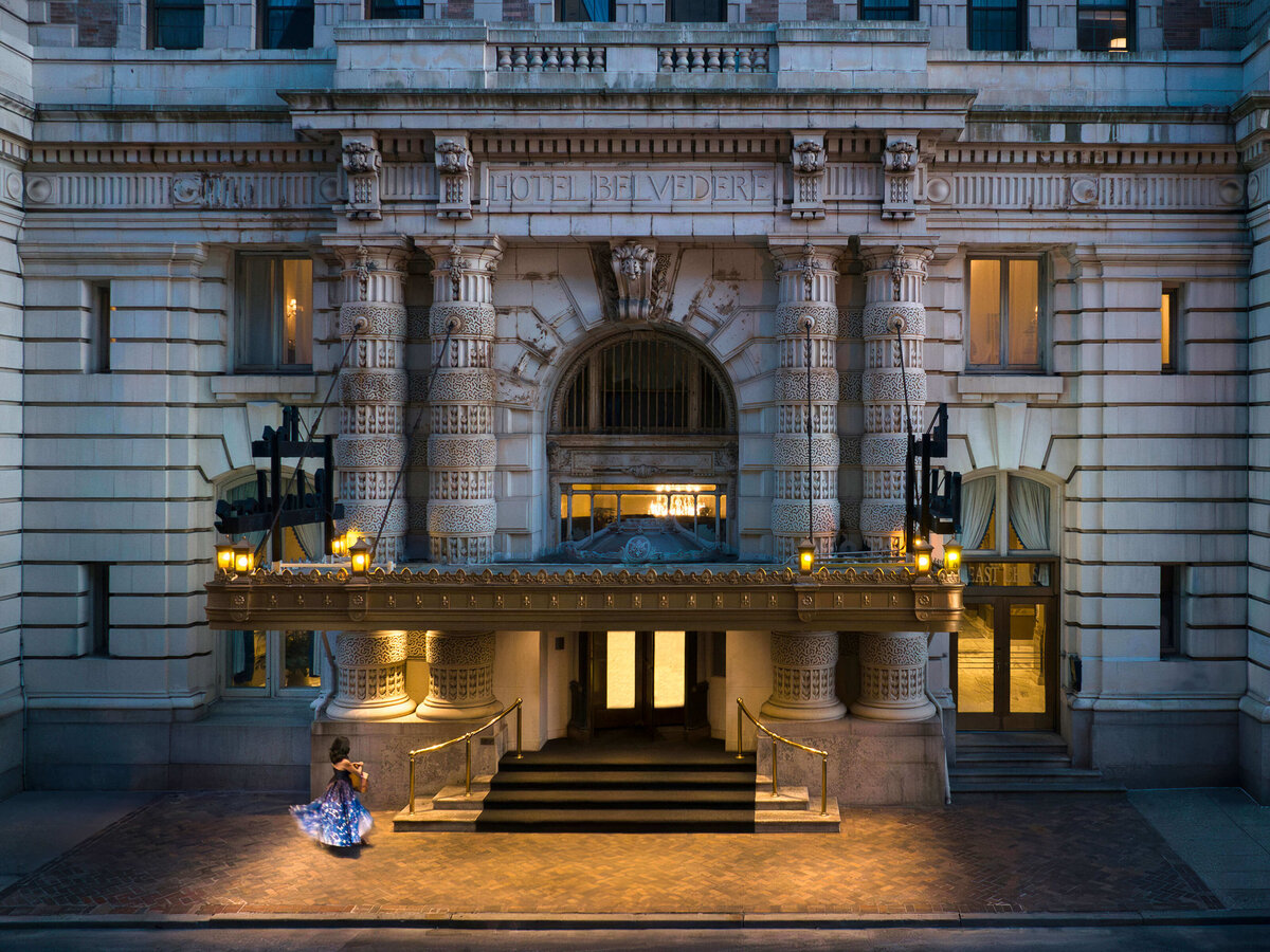 A grand historic hotel entrance with intricate stone carvings and a gold canopy that reads Hotel Belvedere. A woman in a flowing blue dress dances gracefully on the spacious sidewalk in front.