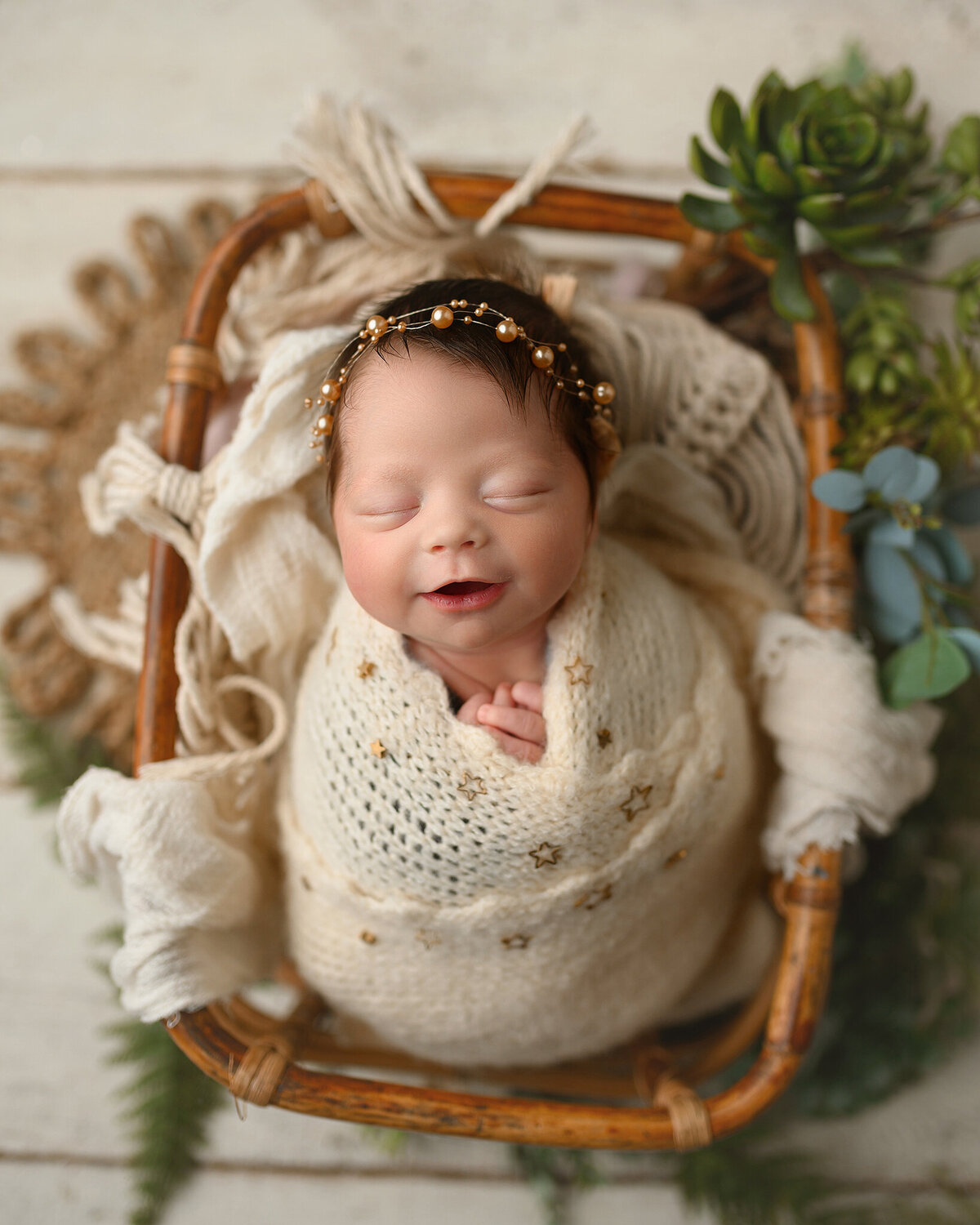 adorable baby girl with dark hair, smiling swaddled in soft yellow wrap, in a bamboo basket with succulents around her