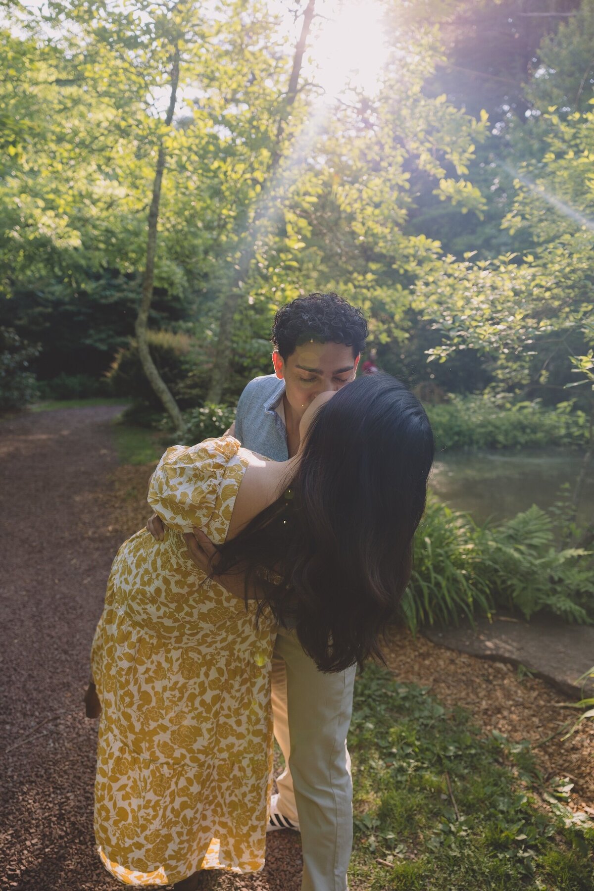 Groom-to-be lifting bride-to-be into a playful hug with sunlight filtering through the trees.