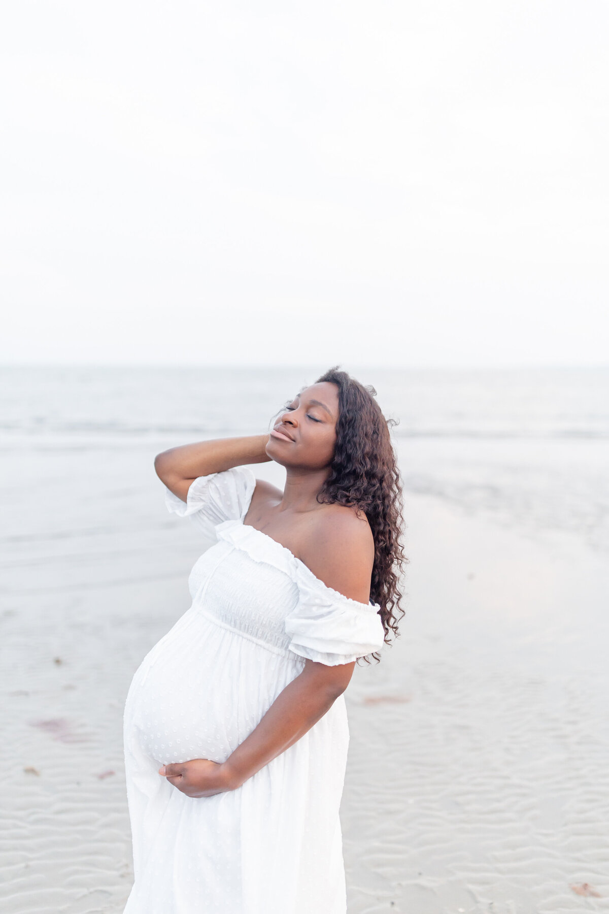 a pregnant mother stands on a beach with her face towards the sun and her eyes closed