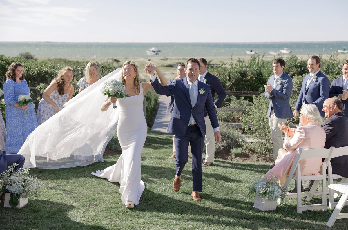 Bride and Groom celebrate as they walk hand in hand down the aisle with view of water behind them.