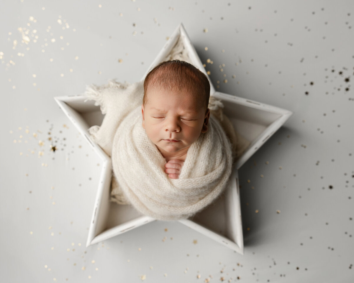 Newborn baby boy wrapped in cream knits and posed inside a white star prop with gold star confetti sprinkled around. Captured by Inland Empire newborn photographer Kaitlyn Dawn Photography.