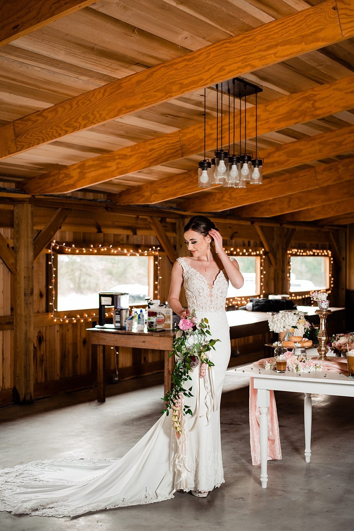 A bride wearing a wedding dress with a plunging neckline, beaded bodice and long sating train stands beside a white farm table with assorted desserts holding a long cascade bouquet of pink peony, king protea, Italian ruscus, a long garland of white roses and long flowing white satin ribbons at Grace Valley Farm.