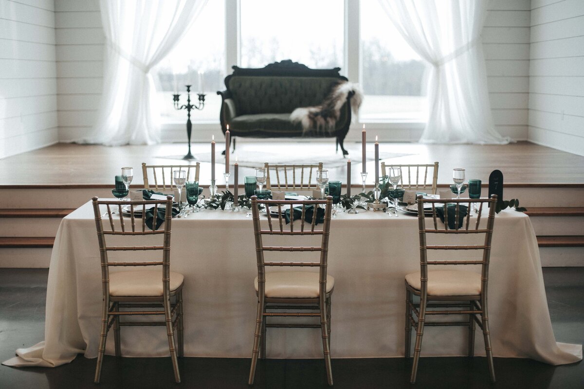 Wedding reception table set with ivory table cloth, and gold and dark green accents on a wedding venue with airy lighting.