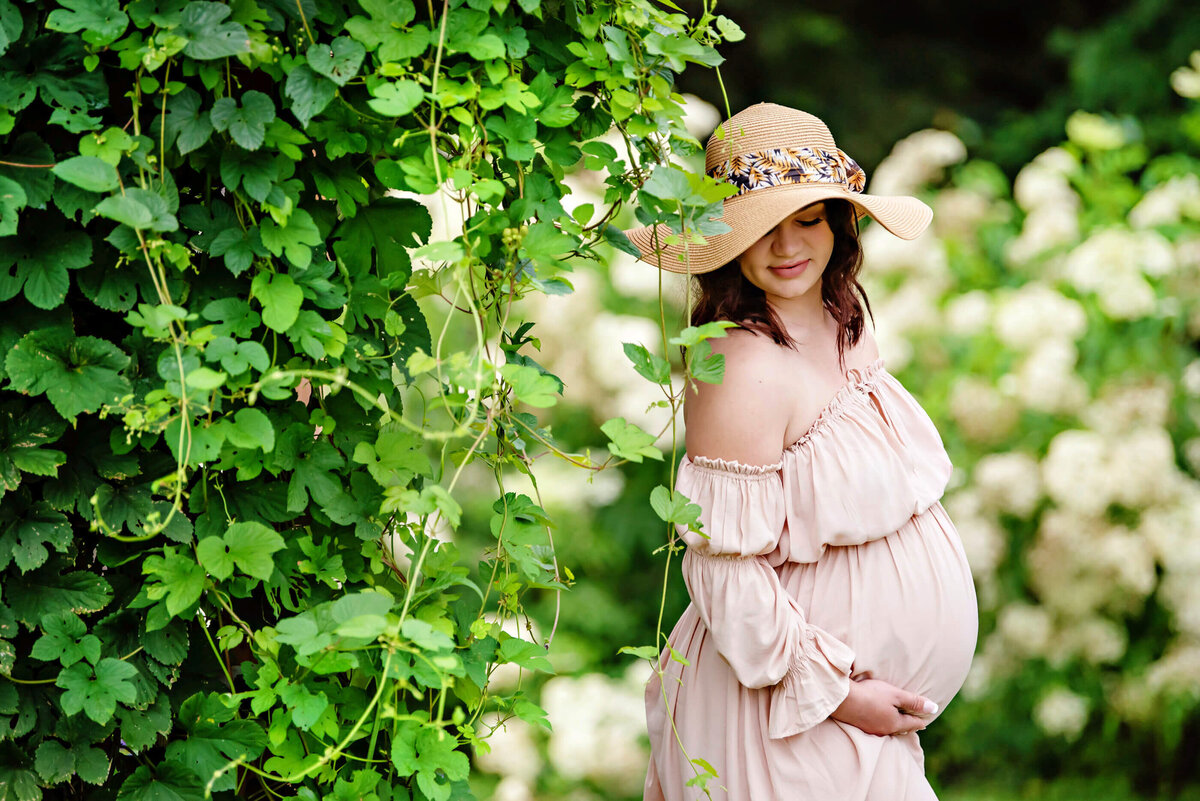 pregnant woman wearing a pink maternity dress standing against some flowers for her outdoor maternity photos