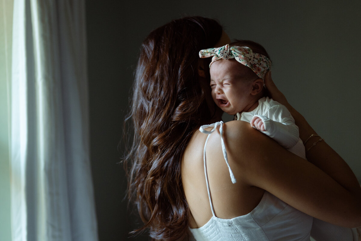 A serene image of a baby resting on the mother’s shoulder, enveloped in soft natural light, captured during a home lifestyle session in Fort Worth.