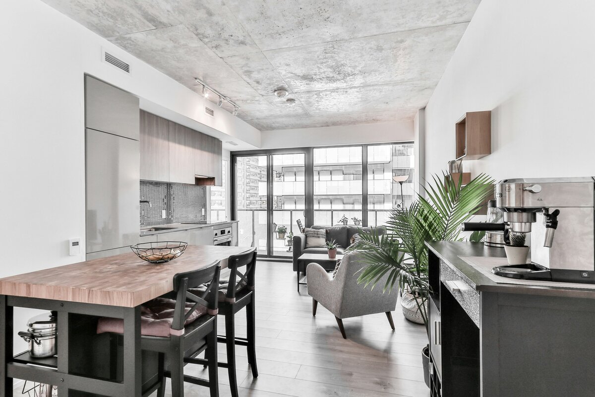A modern, bright apartment interior showcasing an open-concept living space. A concrete ceiling with exposed ductwork gives it an industrial loft feel. In the foreground, a kitchen island with a wooden countertop and black barstools is visible. Beyond, a living area features a dark sofa and a light-colored armchair facing a large window that opens onto a balcony. A potted plant adds a touch of greenery to the foreground. The overall impression is minimalist and contemporary.