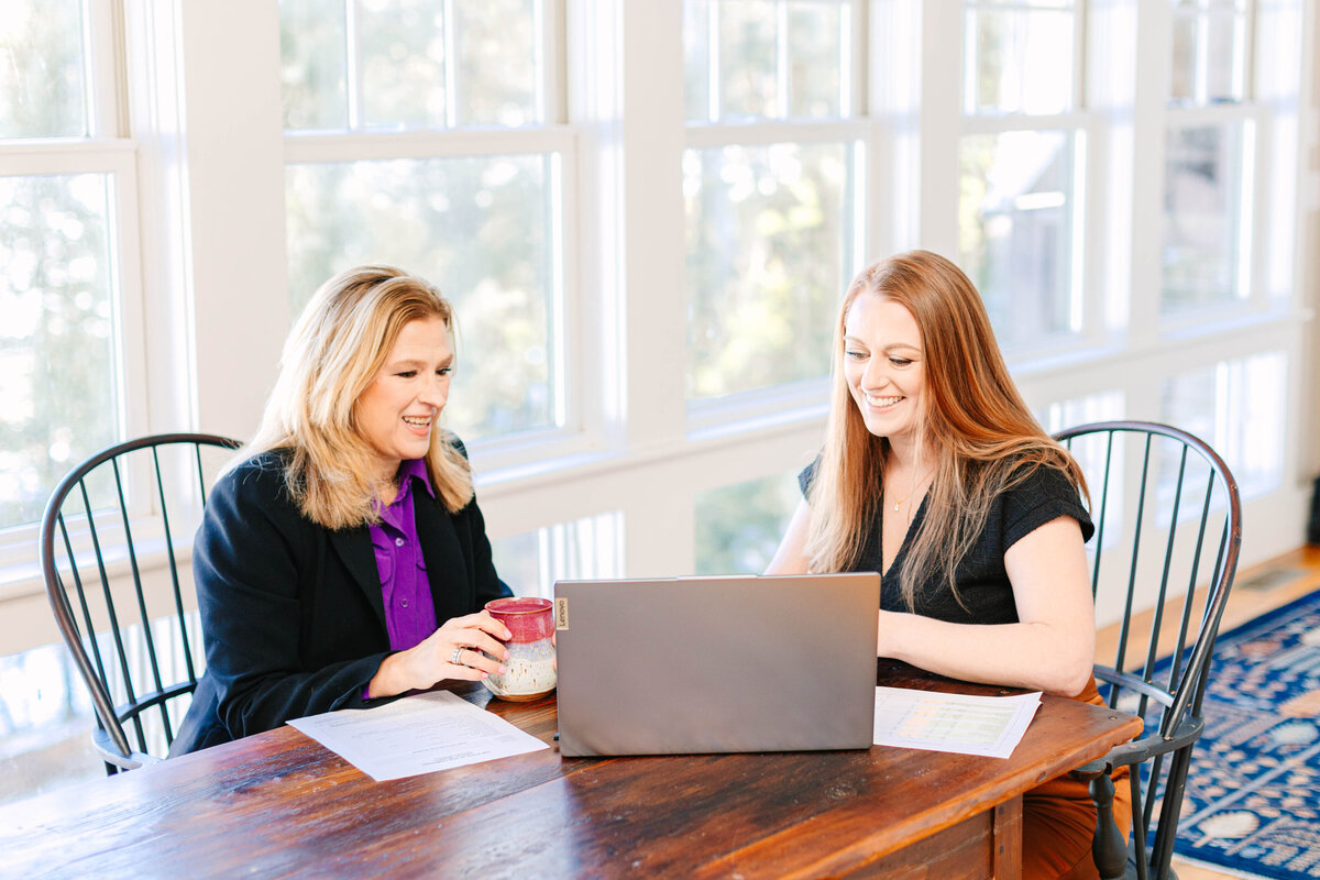 Two professional women sit at a wooden table in a bright office space, smiling as they work on a laptop. One holds a coffee mug, while the other types, with documents spread out on the table