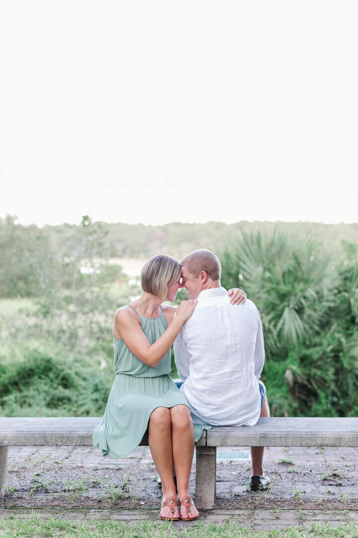 A couple sits on a bench with the marsh behind them, staring into each others eyes. Captured by Arkansas Wedding Photographer, Photography by Karla.