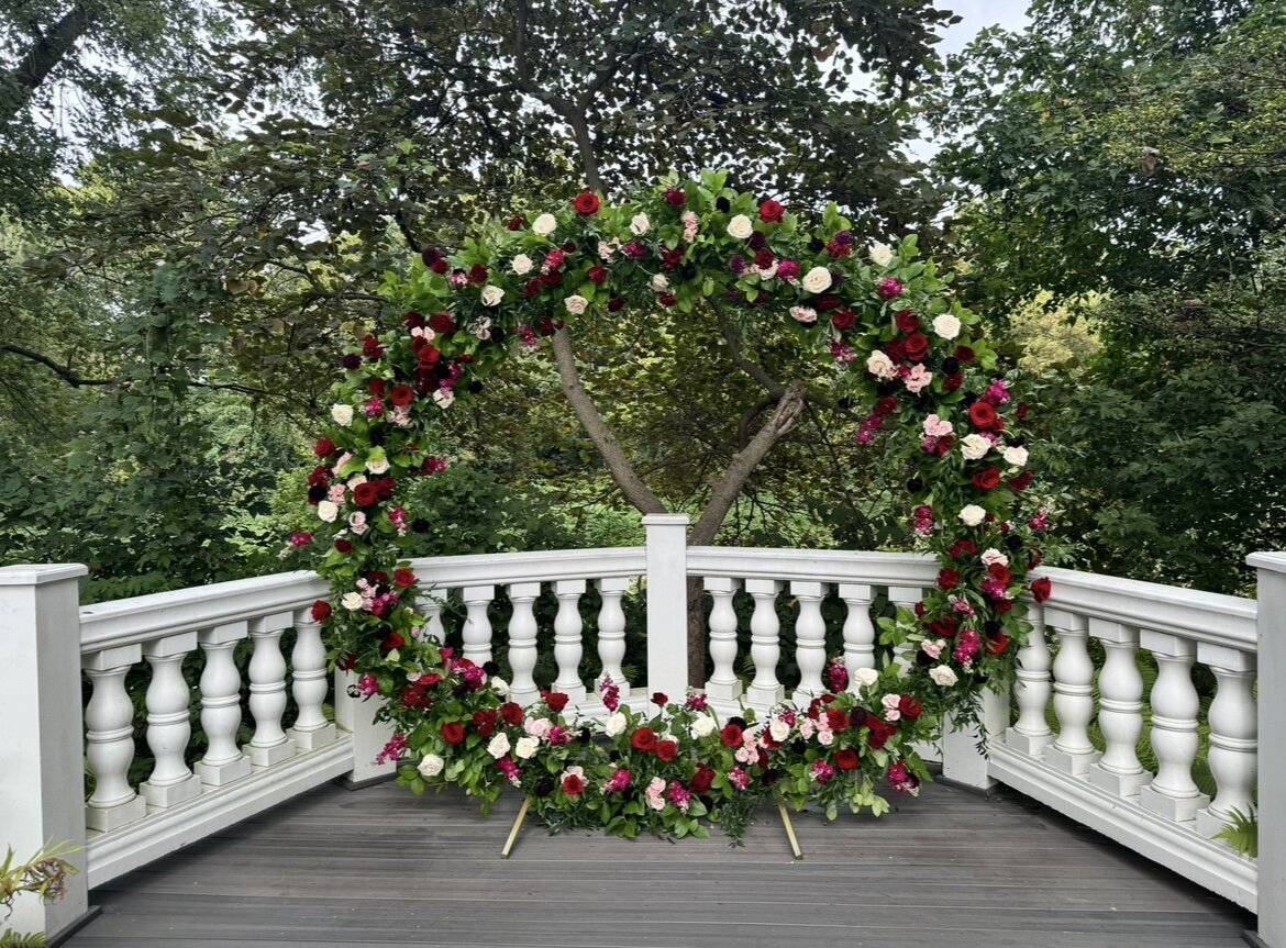 Round floral ceremony arch adorned with red, pink, and blush flowers, set outdoors at the Elkridge Furnace Inn. The arch is framed by lush greenery and a classic white railing, creating a romantic and elegant setting.