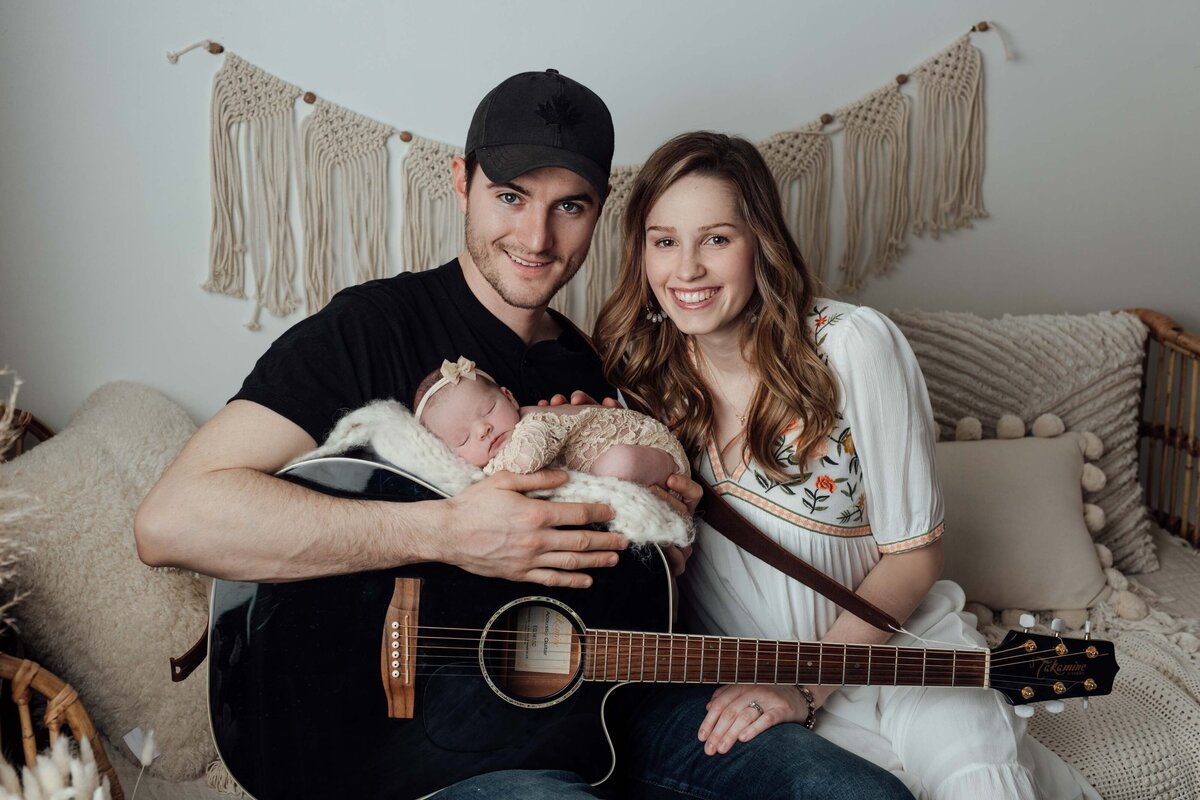 Studio newborn session - Mom and Dad sitting on a couch. Dad has is guitar resting on his lap and the baby is  sleeping on top of the guitar wearing a beige bodysuit and matching headband.