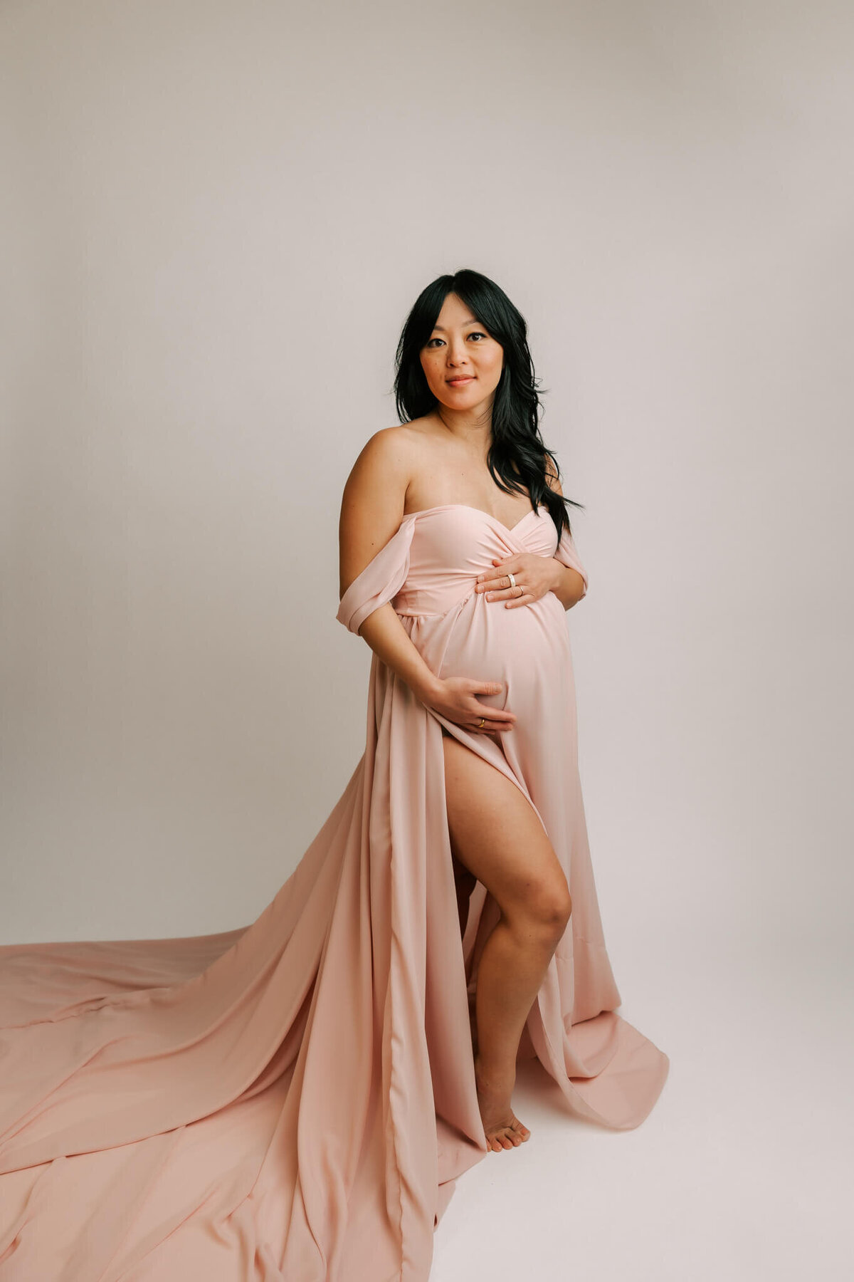 studio maternity portrait of a mom wearing a pink dress and smiling at the camera with her hangs on her belly