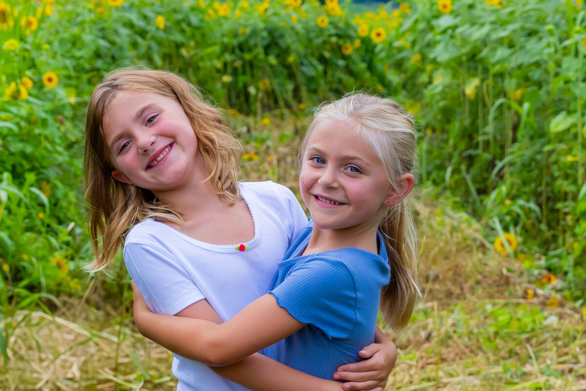 two sisters hugging in a sunflower field smiling at the camera